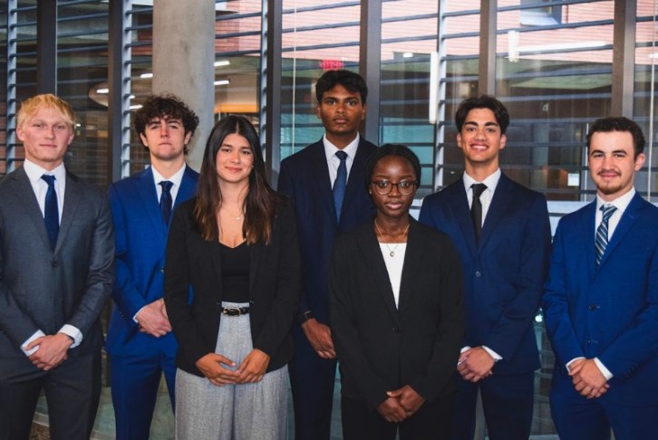 Seven students in business attire standing in front of a glass building