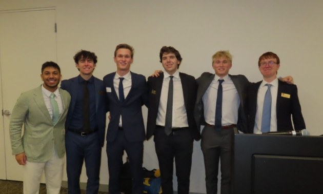 Six students in suits standing together beside a podium